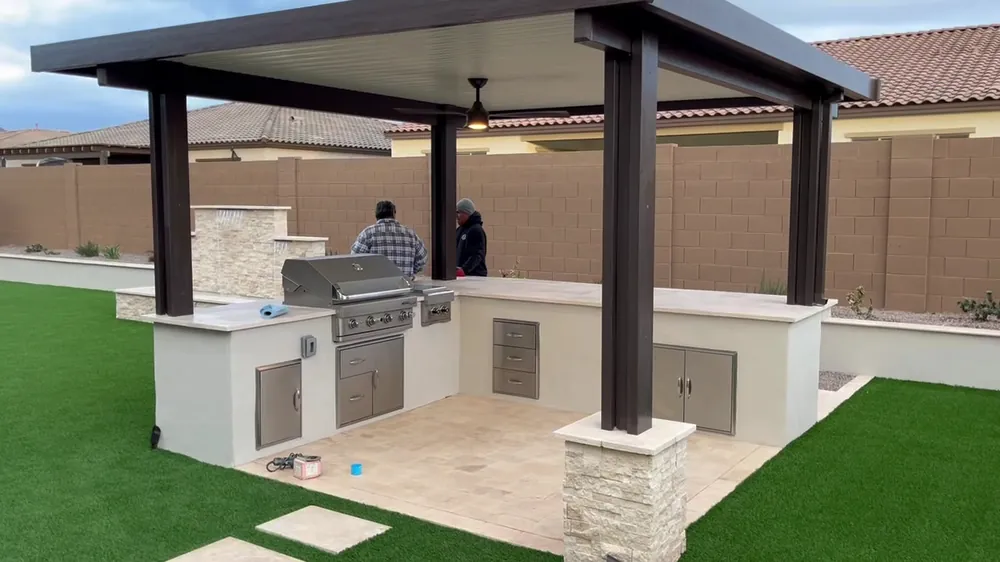 Outdoor kitchen with a pergola, stone-clad columns, a grill and artificial turf, with two crew members at work
