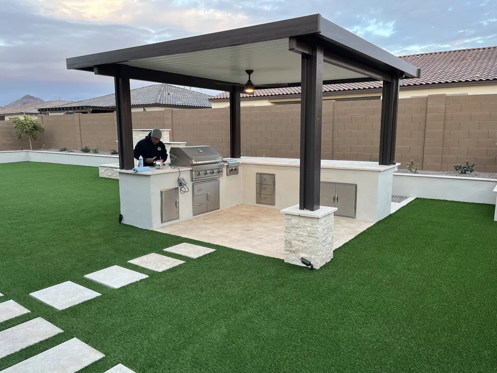 Outdoor kitchen under an aluminum pergola with a built-in grill, stone-clad columns and artificial turf