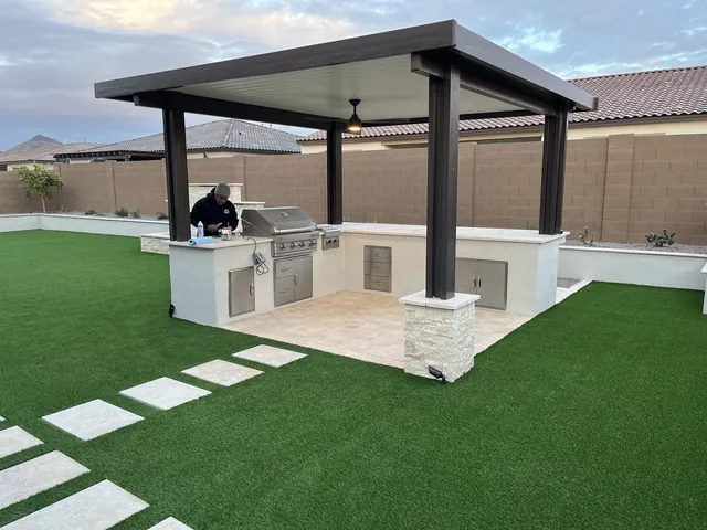 Outdoor kitchen under an aluminum pergola with a built-in grill, stone-clad columns and artificial turf
