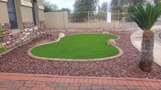 Kidney-shaped artificial turf lawn edged in stone, surrounded by red gravel with a palm beside it